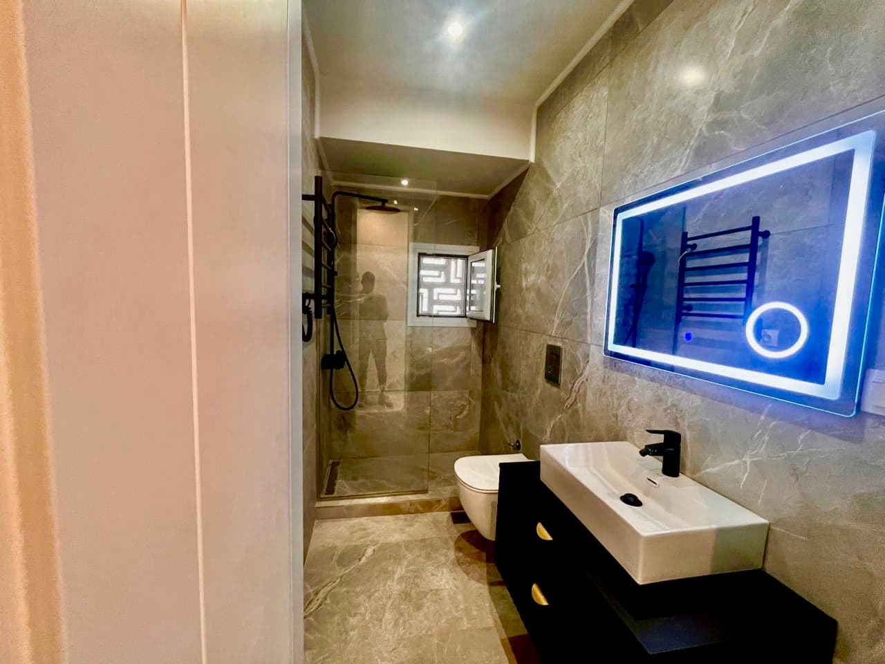 The same bathroom after renovation with grey marble-effect tiles, walk-in shower, wall-hung WC and backlit mirror